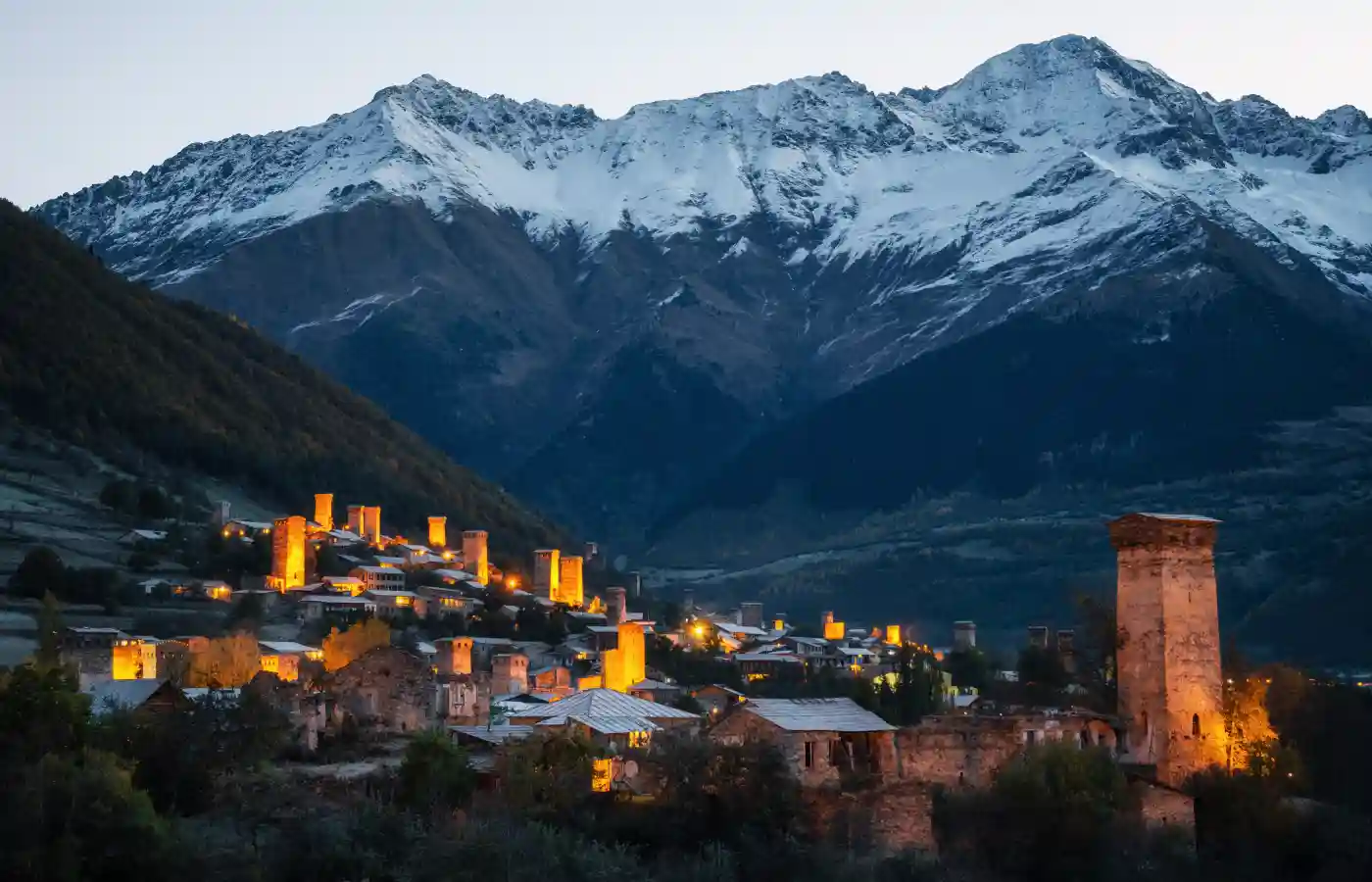 View of the Svanetian towers with night illumination in Mestia village against snowy mountains at sunrise. Upper Svaneti, Georgia.