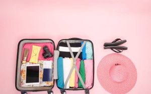 Suitcase packed with travel essentials with a hat and slipper near it.