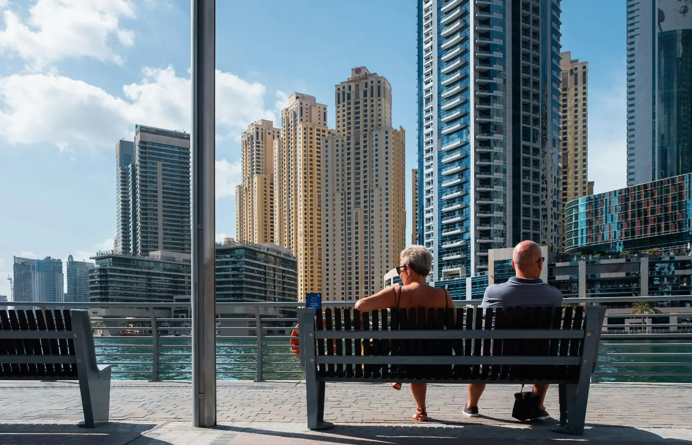 an elderly couple seated on a bench observing the Dubai creek overlooking buildings of Dubai